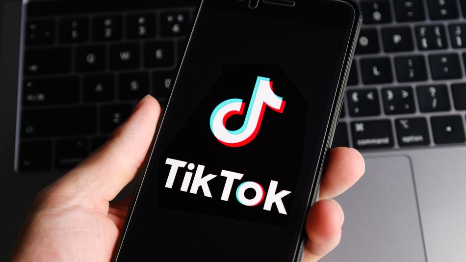 Рука на фоне серого ноутбука держит смартфон с логотипом ТикТок