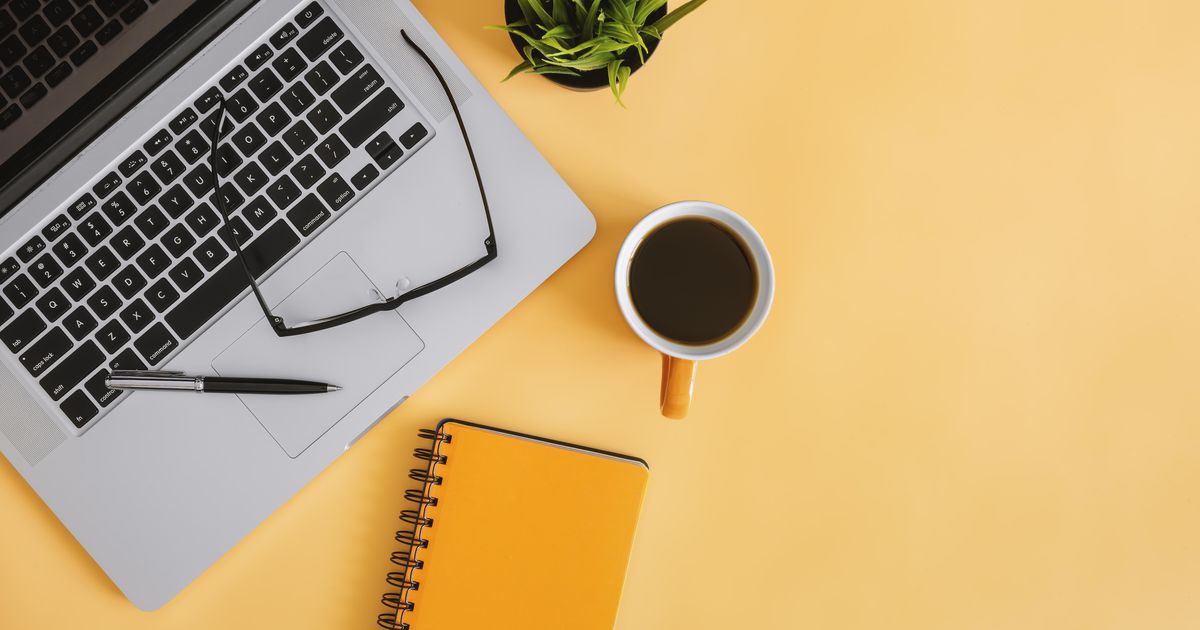 На жёлтом столе стоит ноутбук, лежит оранжевый ежедневник и стоит чашка кофе