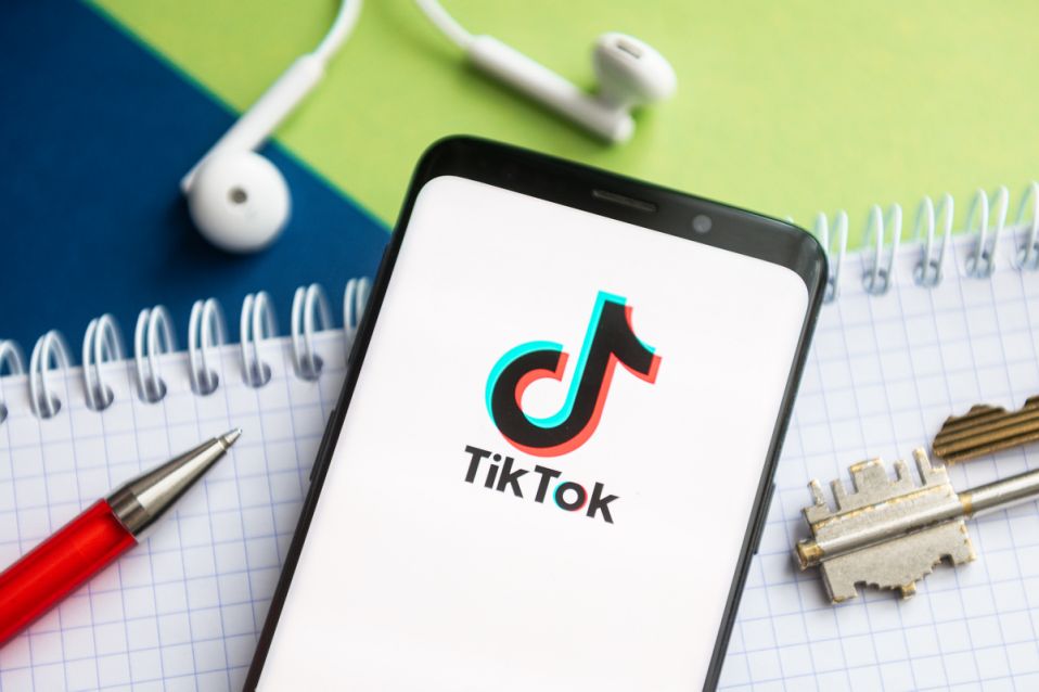 Лого ТикТока на смартфоне, лежащем на тетради рядом с ручкой, ключами и наушниками